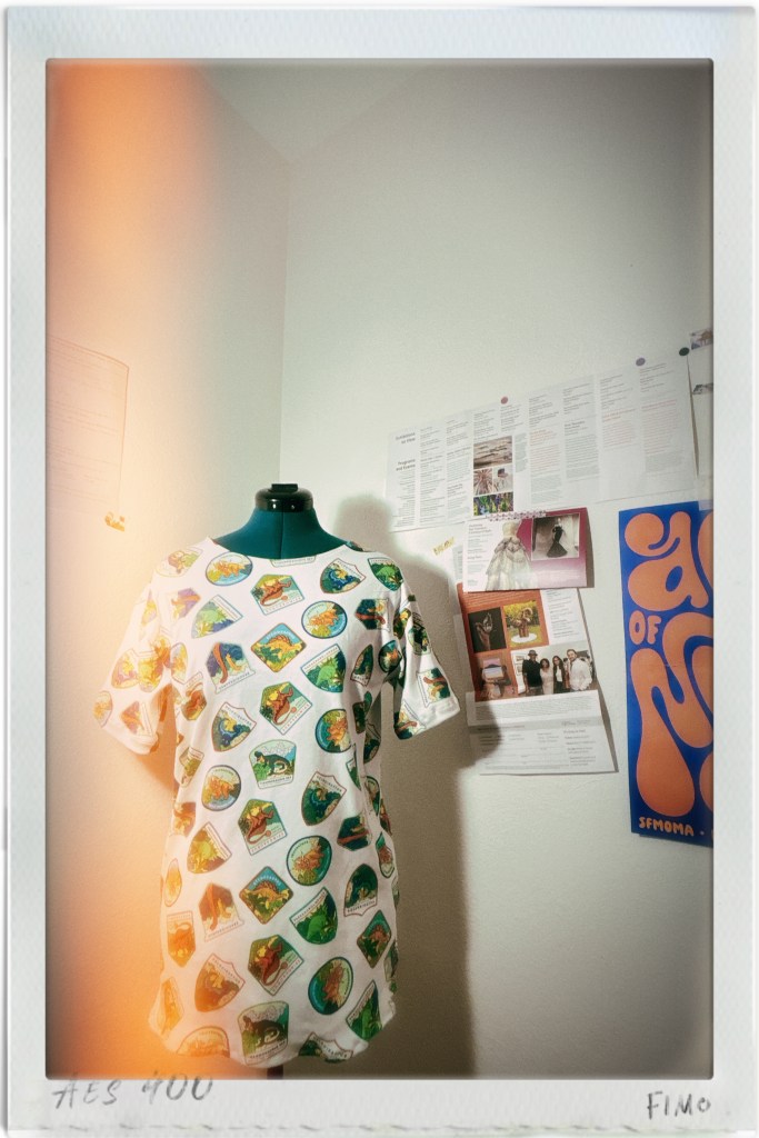 A T-shirt with a colorful dinosaur print displayed on a dress form against a wall with various notices and posters.